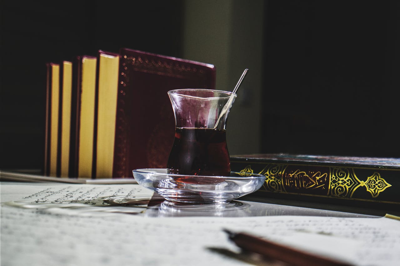 A glass of black tea on a table with ornate books including the Quran, enhancing a scholarly atmosphere.
